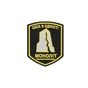 May include: A shield-shaped embroidered patch with a black background and yellow border. It features a white stylized rock formation with a central crack. The text "СИЛА В ЄДНОСТІ" is arched at the top, and "МОНОЛІТ" is printed at the bottom in yellow.