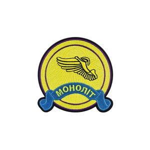 May include: A circular embroidered patch featuring a bright yellow background with a black border. In the center, a stylized black wing graphic is displayed. Below the wing, a blue ribbon banner contains the yellow Cyrillic text МОНОЛІТ.