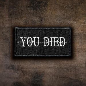 May include: A rectangular black fabric patch featuring the white embroidered text YOU DIED. The text is bisected by a horizontal white line that mimics the appearance of a flatline heart rate monitor signal, centred on the patch.