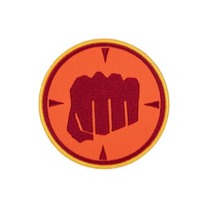 Könnte beinhalten: Ein kreisförmiger gestickter Aufnäher mit einer dunkelroten Faustsilhouette auf leuchtend orangefarbenem Hintergrund. Das Design ist von einem dünnen dunkelroten Kreisrand und einem gelben Außenrand umgeben, mit vier kleinen dreieckigen Markierungen.