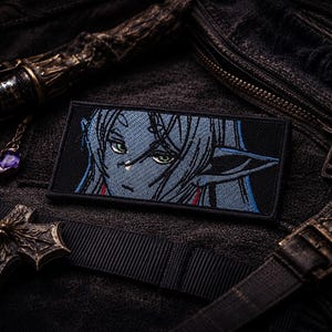 May include: A rectangular embroidered patch featuring a stylized illustration of a blue-skinned character with pointed ears and green eyes. The character has long hair and is depicted from the chest up against a solid black background.