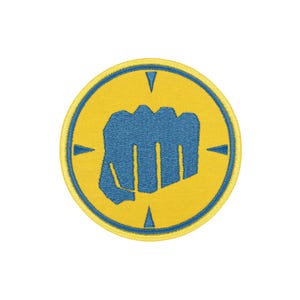 Op de afbeelding: Een ronde geborduurde patch met een gele achtergrond en een blauwe rand. In het midden is een blauwe vuist afgebeeld. Het ontwerp bevat vier blauwe driehoekige markeringen die vanaf de rand naar binnen wijzen, geplaatst aan de boven-, onder-, linker- en rechterkant.