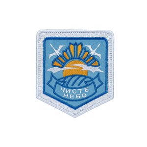 May include: A shield-shaped embroidered patch with a light blue background, featuring a yellow sun rising over a field, two white birds in flight, and a blue banner at the bottom with the white Cyrillic text "ЧИСТЕ НЕБО" stitched in the center.