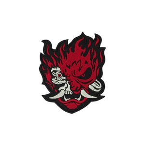 Op de afbeelding: Een geborduurde patch met een gestileerd rood en zwart demonenmaskerontwerp. De afbeelding toont een gezicht dat half uit een schedel en half uit een masker bestaat, met scherpe witte hoektanden, omringd door vlamvormen in felrood garen.