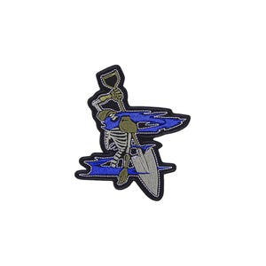May include: An embroidered patch featuring a skeleton figure holding a shovel, surrounded by stylized blue flame-like shapes. The design is outlined in black thread, with the skeleton rendered in white and grey, and the shovel in metallic grey tones.