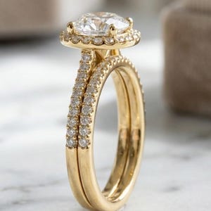 Peut inclure: Une bague en or composée de deux anneaux. L'anneau principal présente une grande pierre ronde à facettes transparentes entourée d'un halo de petites pierres. Les deux anneaux sont ornés d'un pavage de minuscules pierres scintillantes.