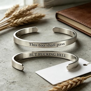Puede incluir: Dos brazaletes abiertos de metal plateado. El brazalete superior tiene grabado el texto "This too shall pass" en la superficie exterior, mientras que el brazalete inferior presenta el texto grabado "BUT FUCKING HELL" en la superficie interior.