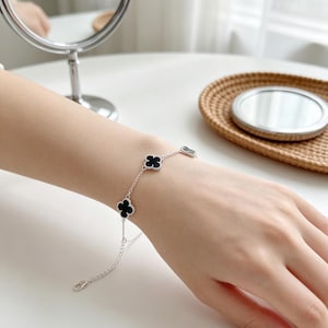 Puede incluir: Una pulsera de cadena plateada con múltiples dijes negros en forma de trébol. La pulsera tiene un diseño de eslabones delicados e incluye una cadena de extensión ajustable con un cierre de mosquetón, mostrando un motivo de trébol.
