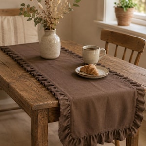 Op de afbeelding: Een rechthoekige bruine tafelloper met een geplooide randafwerking langs de gehele omtrek. De stof heeft een effen, matte textuur en ligt plat over het oppervlak van een houten tafel, wat zorgt voor een decoratieve rand voor het tafelblad.
