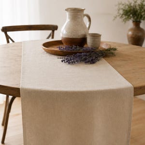 May include: A rectangular beige table runner made of a woven fabric texture draped over a round wooden table. The runner features a clean hemmed edge and a neutral solid colour, providing a flat surface for a wooden tray with a ceramic pitcher and lavender.