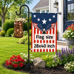 May include: A black metal garden flag stand holding a rectangular flag with a bald eagle graphic, stars, and red and white stripes. The flag features the text "Flag size 28x40 inch". A copper-colored metal lantern with floral cutouts hangs from the stand.