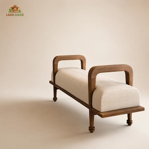 May include: A wooden bench featuring a long, rectangular, off-white upholstered seat cushion. The frame is constructed from dark brown wood with two arched armrests on each side and four turned legs. The logo LAKDI GOCHA is visible in the corner.