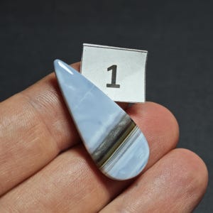May include: A teardrop-shaped cabochon gemstone featuring a smooth, light blue surface with a distinct horizontal band of dark gray and white mineral inclusions. A small white paper tag with the black printed number 1 is positioned behind it.