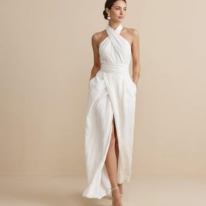 May include: A floor-length white maxi dress featuring a halter neckline with a crossover bodice design, a fitted waistband, and a skirt with a high front slit. The garment includes side pockets and is constructed from a solid white fabric.