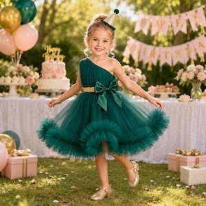 May include: A dark green one-shoulder dress featuring a layered tulle skirt with a ruffled hem, a gold sequined waistband, and a large matching green bow at the waist. The dress is worn with a small gold party hat and gold ballet flats.