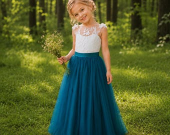 May include: A floor-length dress featuring a white sleeveless bodice with floral lace detailing and a solid teal blue satin waistband. The skirt is made of layered teal blue tulle, creating a full silhouette. The dress is held with a small bouquet.