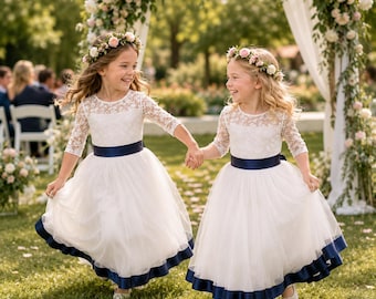 May include: Two white dresses featuring lace bodices with three-quarter length sleeves, round necklines, and full tulle skirts. Each dress is accented with a dark navy blue satin ribbon at the waist and a matching navy blue ribbon trim at the hem.