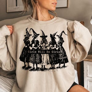 Pu&ograve; includere: Una felpa girocollo color beige con una stampa grafica nera di cinque figure che indossano cappelli da strega a punta e abiti lunghi, in piedi in cerchio con le braccia intrecciate. Il testo "Girls Will Be Girls" &egrave; stampato in nero.