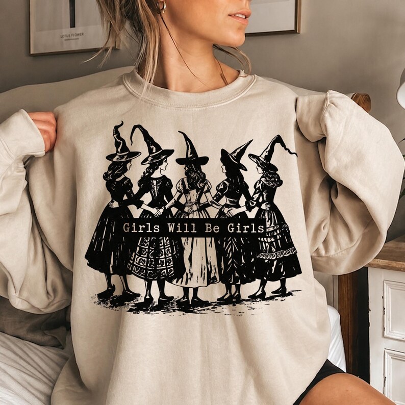 Pu&ograve; includere: Una felpa girocollo color beige con una stampa grafica nera di cinque figure che indossano cappelli da strega a punta e abiti lunghi, in piedi in cerchio con le braccia intrecciate. Il testo "Girls Will Be Girls" &egrave; stampato in nero.