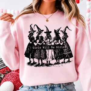 Pu&ograve; includere: Una felpa girocollo rosa chiaro con una stampa grafica nera di cinque figure che indossano cappelli da strega a punta e abiti lunghi, in piedi in cerchio tenendosi per mano. Il testo "Girls Will Be Girls" &egrave; stampato in bianco su una barra nera.