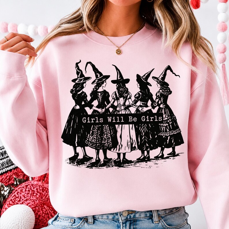 Pu&ograve; includere: Una felpa girocollo rosa chiaro con una stampa grafica nera di cinque figure che indossano cappelli da strega a punta e abiti lunghi, in piedi in cerchio tenendosi per mano. Il testo "Girls Will Be Girls" &egrave; stampato in bianco su una barra nera.