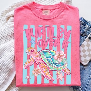 May include: A coral pink t-shirt featuring a graphic of a sea turtle with a teal and pink shell, a pink hibiscus flower, and two stars. The text "TURTLEY OVER IT" is printed in pink above the turtle, set against a background of vertical light blue stripes.