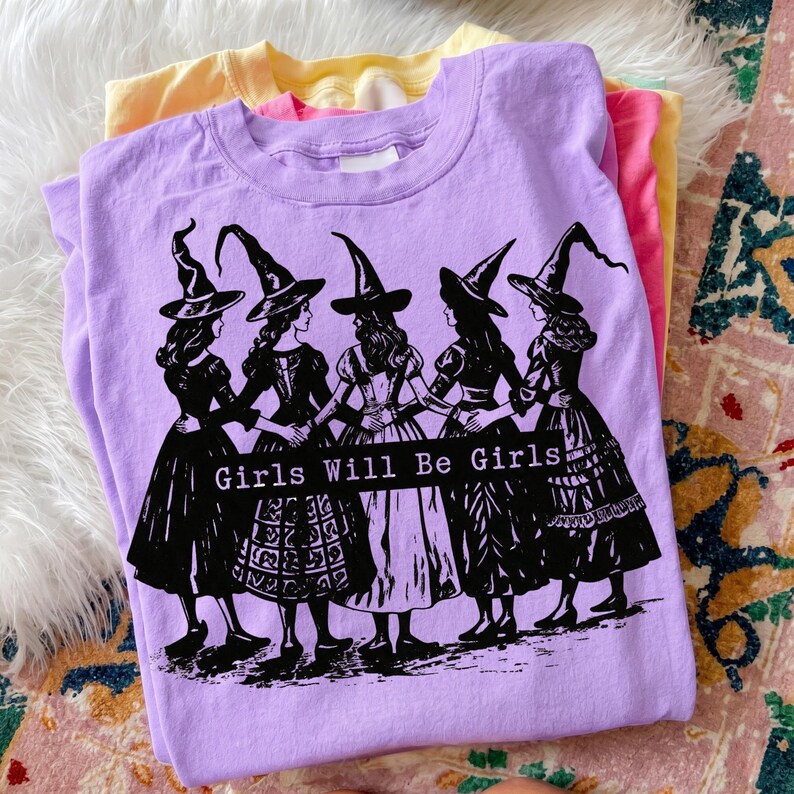 Pu&ograve; includere: Una t-shirt a maniche corte color lavanda con una stampa grafica nera di cinque figure che indossano cappelli da strega a punta e abiti lunghi, in piedi in cerchio tenendosi per mano. Il testo "Girls Will Be Girls" &egrave; stampato in bianco su una barra nera.