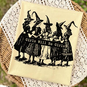 Pu&ograve; includere: Una t-shirt giallo chiaro con una stampa grafica nera di cinque figure che indossano cappelli a punta e abiti lunghi, in piedi in cerchio e tenendosi per mano. Un banner nero centrale contiene il testo Girls Will Be Girls.