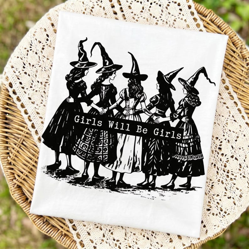 Pu&ograve; includere: Una t-shirt bianca con una stampa grafica nera di cinque figure che indossano cappelli da strega a punta e abiti lunghi, in piedi in cerchio mentre si tengono per mano. Al centro dell'illustrazione, il testo recita, Girls Will Be Girls.