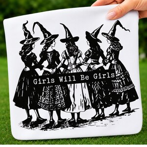 Pu&ograve; includere: Una pochette in tessuto bianco con un'illustrazione grafica nera di cinque figure che indossano cappelli da strega a punta e abiti lunghi, in piedi in cerchio tenendosi per mano. Un banner nero orizzontale al centro mostra Girls Will Be Girls.