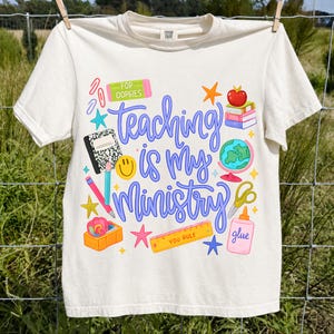 May include: An off-white short-sleeve t-shirt featuring a colorful graphic with the text "Teaching is my Ministry" in blue script. The design includes school supplies like a globe, books, a ruler labeled "YOU RULE," glue, and a smiley face.