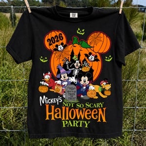 以下が含まれることがあります： ハロウィンの仮装をしたディズニーキャラクター（ミッキーマウス、ミニーマウス、ドナルドダック、グーフィー、プルート）のグラフィックが描かれた黒いTシャツ。デザインにはカボチャ、「Trick or Treat」と書かれた墓石、そして「Mickey's Not So Scary Halloween Party 2026」というテキストが含まれています。