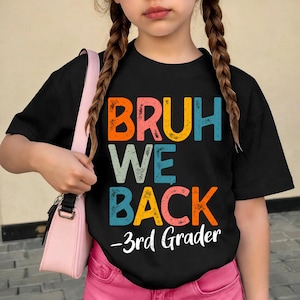 May include: A black short-sleeved t-shirt featuring the distressed, multi-colored text "BRUH WE BACK" in orange, yellow, teal, and pink, with the white script text "3rd Grader" underneath. The shirt is paired with a light pink shoulder bag.