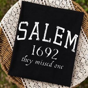 May include: A black t-shirt featuring white distressed lettering that reads SALEM 1692 they missed one. The word SALEM is printed in a large, bold, block-style font, while 1692 and the phrase they missed one appear in a smaller, serif font.