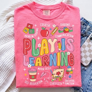 Peut inclure: Un t-shirt rose corail avec un motif coloré portant le texte PLAY IS LEARNING. Le design comprend des illustrations de blocs alphabétiques, une pomme, un crayon, des briques de construction, une tasse de café, un chevalet et des étoiles.
