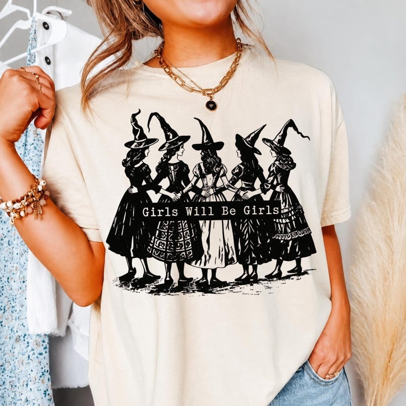 Pu&ograve; includere: Una t-shirt color crema con una stampa grafica nera di cinque figure che indossano cappelli da strega a punta e abiti lunghi, in piedi in cerchio con le braccia intrecciate. Il testo "Girls Will Be Girls" &egrave; stampato in una barra nera.