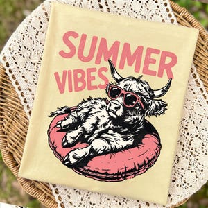 May include: A light yellow t-shirt featuring a graphic of a Highland cow wearing pink sunglasses while lounging on a pink inflatable pool float. Above the cow, the text "SUMMER VIBES" is printed in a bold, distressed pink font. The design is centered.