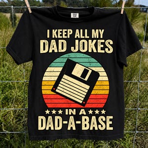 Puede incluir: Una camiseta negra de manga corta con un gráfico de un disquete sobre un círculo de atardecer retro. El texto dice I KEEP ALL MY DAD JOKES IN A DAD-A-BASE con estrellas flanqueando la línea inferior. El diseño utiliza tonos amarillos, naranjas y rojos.