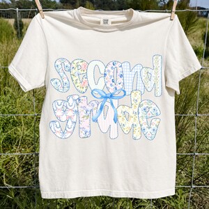 Può includere: Una t-shirt a maniche corte color crema con la scritta "second grade" in un grande carattere a motivi multipli. Le lettere sono riempite con vari disegni floreali, a quadretti e a righe in blu, rosa e giallo, accentuati da un fiocco blu.