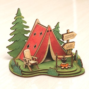 May include: A layered wooden craft featuring a red tent with diamond cutouts, a light-coloured chair, and a campfire with a pot. Green trees and bushes surround the scene, with two wooden signs pointing toward HIKING and LAKE.