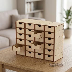 Può includere: Un mobiletto in legno chiaro con ventiquattro piccoli cassetti disposti a griglia. Ogni cassetto presenta un'apertura semicircolare come maniglia. L'unità è costruita con giunzioni visibili e poggia su una superficie in legno.