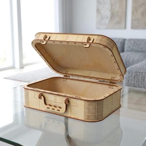 May include: A light-coloured wooden suitcase with an open lid, featuring a laser-cut design with visible interlocking joints, a rectangular frame, and a decorative handle on the front panel. The structure is made of thin plywood sheets.