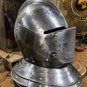 May include: A steel close helmet featuring a rounded skull, a hinged visor with a horizontal eye slit, and a series of small ventilation holes on the right side. The helmet includes a gorget base with metal rivets and a weathered, textured finish.