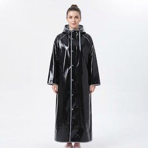 Puede incluir: Un impermeable largo, negro y brillante, con capucha con cordones ajustables, mangas largas y cierre frontal de botones. La prenda presenta ribetes de color gris claro a lo largo de la capucha, la parte delantera y los puños de las mangas.