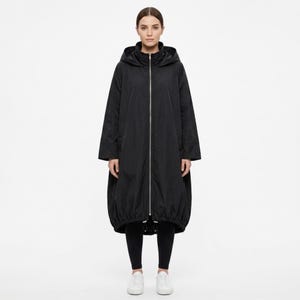 May include: A black long-sleeve hooded coat featuring a full-length silver-tone front zipper, a high collar, and a gathered, elasticized hemline that creates a rounded, voluminous silhouette. The coat is worn over black leggings and white sneakers.