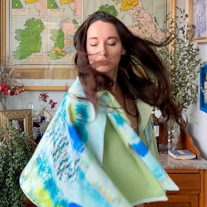May include: A woman wearing a light green top and blue jeans is standing in front of a wooden cabinet. She is wearing a blue, green, and yellow patterned shawl. The shawl is draped over her shoulders and is flowing in the wind.