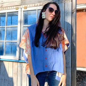 K&ouml;nnte beinhalten: Eine Frau tr&auml;gt ein blaues Hemd mit Kn&ouml;pfen und einem gemusterten Design an den &Auml;rmeln. Sie tr&auml;gt Jeans und hat langes dunkles Haar. Sie tr&auml;gt gro&szlig;e wei&szlig;e Ohrringe und eine Sonnenbrille.