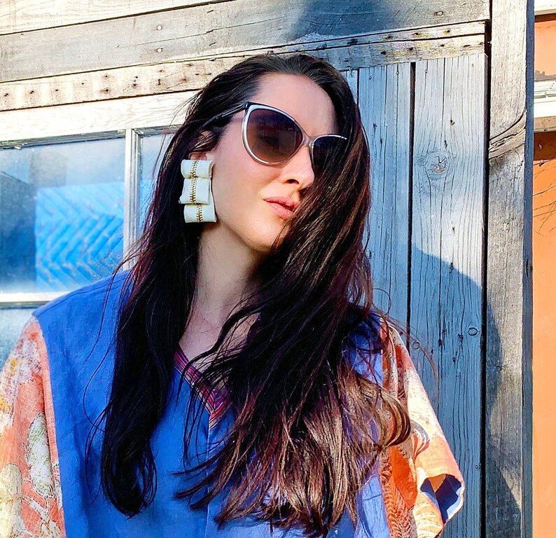 K&ouml;nnte beinhalten: Eine Frau mit langen dunklen Haaren, die ein blau-orangefarbenes gemustertes Hemd und gro&szlig;e wei&szlig;e Statement-Ohrringe tr&auml;gt. Sie tr&auml;gt eine Cat-Eye-Sonnenbrille.
