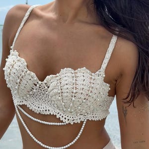 May include: A cream-colored crochet bralette featuring a shell-inspired design with scalloped edges and delicate white pearl embellishments. The garment includes two draped strands of white pearls hanging from the lower edge of the bodice.