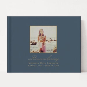 May include: A blue book with a gold frame around a photo of a young woman standing on a beach. The book says "Remembering Virginia Hope Lawrence March 2, 1959 - June 29, 2024".